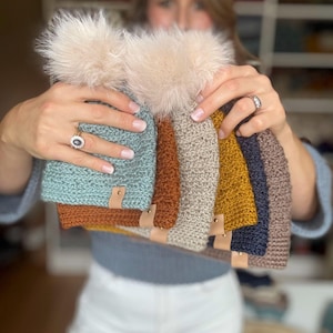 May include: A collection of handmade knit beanies in various colors, including teal, rust, tan, mustard, navy, and brown. Each beanie features a faux fur pom-pom and a small leather tag. The beanies are displayed in a person's hands.