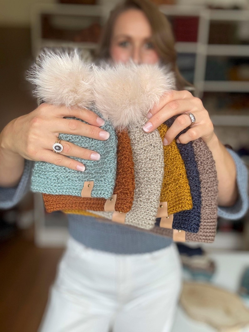 May include: A collection of handmade knit beanies in various colors, including teal, rust, tan, mustard, navy, and brown. Each beanie features a faux fur pom-pom and a small leather tag. The beanies are displayed in a person's hands.