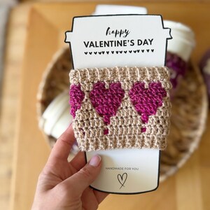 Op de afbeelding: Een handgemaakte Valentijnsdag cadeau set met een beige gehaakte koffiebekerhoes met drie magenta harten. De hoes is gewikkeld rond een witte papieren beker-vormige kaart met de tekst "happy VALENTINE'S DAY" en "HANDMADE FOR YOU".
