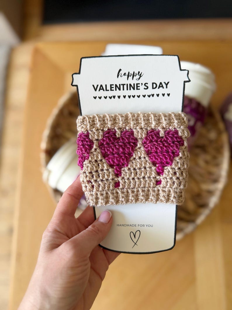 Op de afbeelding: Een handgemaakte Valentijnsdag cadeau set met een beige gehaakte koffiebekerhoes met drie magenta harten. De hoes is gewikkeld rond een witte papieren beker-vormige kaart met de tekst "happy VALENTINE'S DAY" en "HANDMADE FOR YOU".