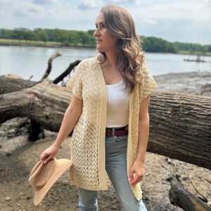 May include: A cream-colored, open-front crochet cardigan with short sleeves. The cardigan features a geometric pattern and is worn over a white top and blue jeans. A straw hat is held in the hand. The background includes a river and trees.