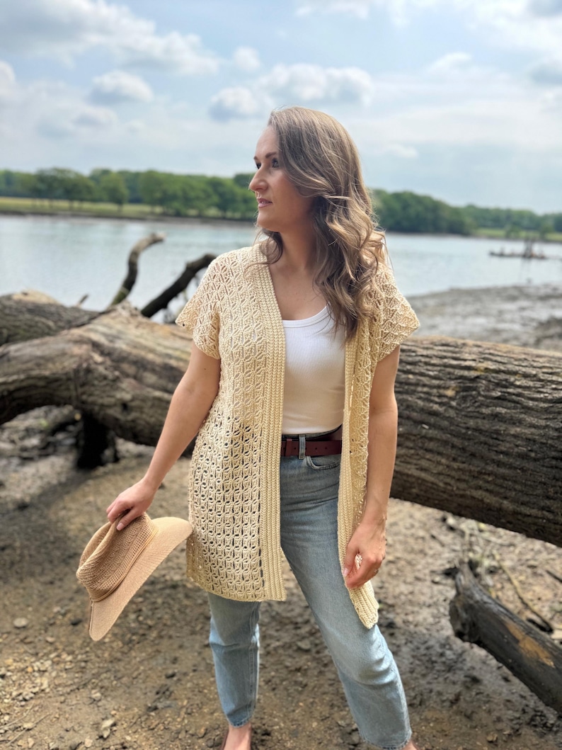 May include: A cream-colored, open-front crochet cardigan with short sleeves. The cardigan features a geometric pattern and is worn over a white top and blue jeans. A straw hat is held in the hand. The background includes a river and trees.