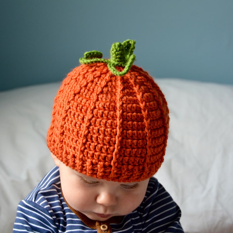 May include: A baby wearing a crocheted pumpkin hat with a green stem. The hat is orange and has a textured surface. The baby is wearing a blue and white striped shirt.