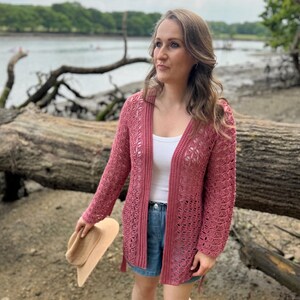 May include: A dusty rose open-front cardigan with a delicate, open-weave design. The cardigan has long sleeves and is worn over a white top and denim shorts. A beige sun hat is held in the hand. The background features a natural outdoor setting.