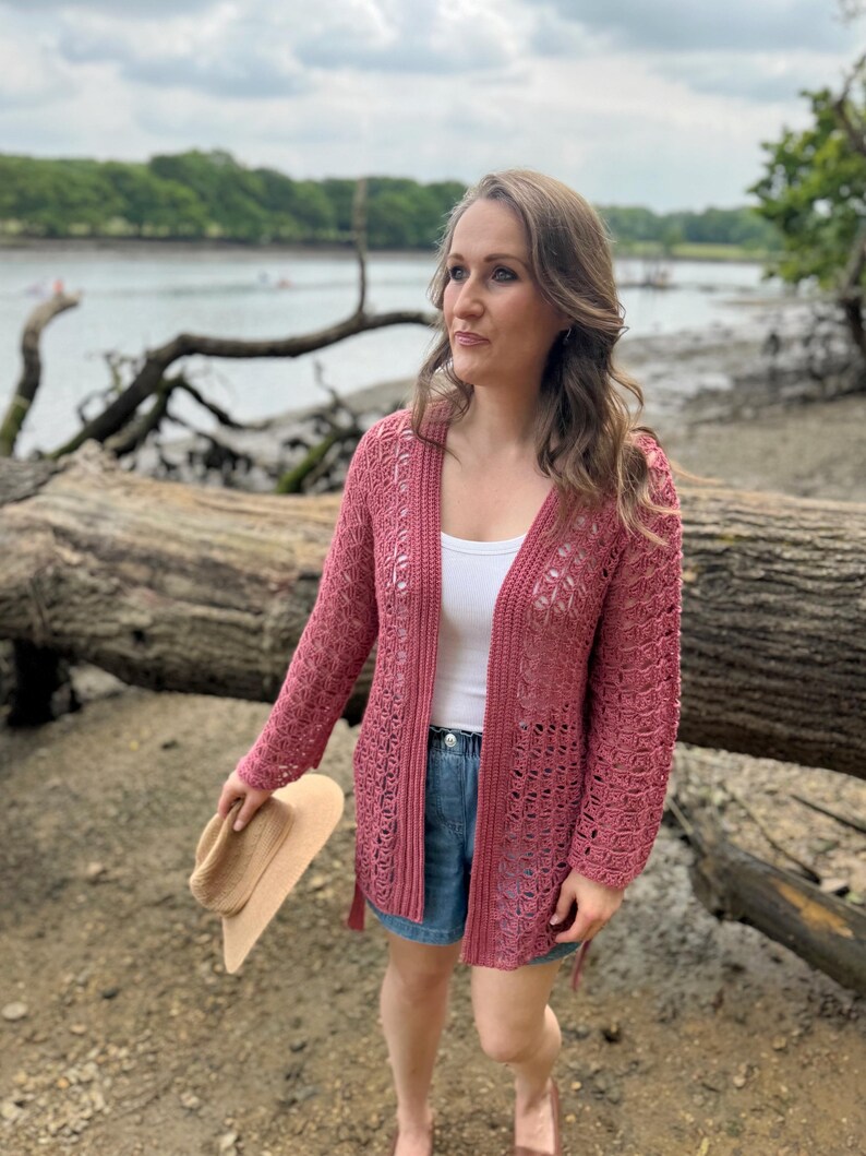 May include: A dusty rose open-front cardigan with a delicate, open-weave design. The cardigan has long sleeves and is worn over a white top and denim shorts. A beige sun hat is held in the hand. The background features a natural outdoor setting.