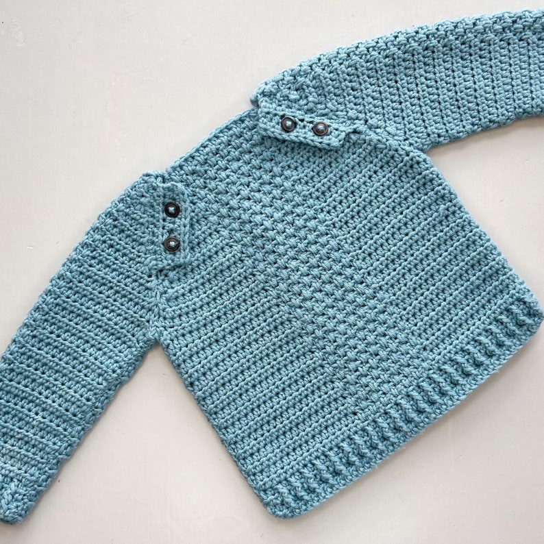 May include: A light blue crocheted sweater with a button closure at the neck. The sweater has a textured stitch pattern.