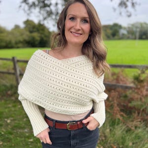 May include: Cream-colored, off-the-shoulder crochet wrap with long sleeves. The wrap is paired with a brown leather belt and dark wash jeans. The background features a green field and a wooden fence.