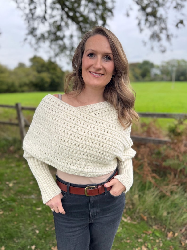 May include: Cream-colored, off-the-shoulder crochet wrap with long sleeves. The wrap is paired with a brown leather belt and dark wash jeans. The background features a green field and a wooden fence.