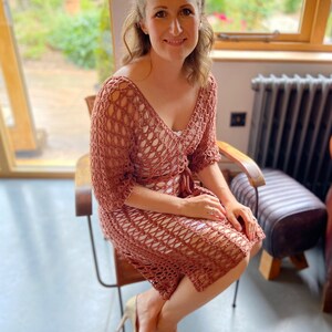 Puede incluir: Una mujer lleva un vestido de ganchillo de color marr&oacute;n claro. El vestido tiene un corte holgado y un delicado patr&oacute;n de calado. Est&aacute; sentada en una silla con estructura de madera y asiento de cuero negro.