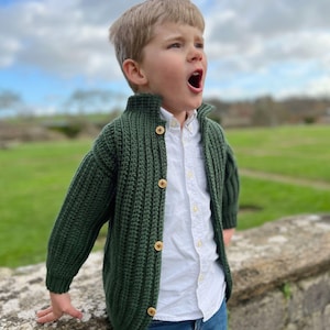 K&ouml;nnte beinhalten: Ein kleiner Junge tr&auml;gt einen gr&uuml;nen Strickcardigan mit Holzkn&ouml;pfen, ein wei&szlig;es Hemd mit Kragen und eine blaue Jeans. Er steht im Freien und schaut nach oben.