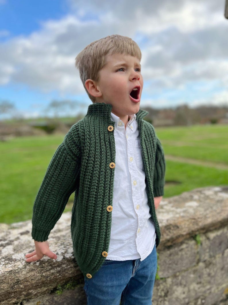 K&ouml;nnte beinhalten: Ein kleiner Junge tr&auml;gt einen gr&uuml;nen Strickcardigan mit Holzkn&ouml;pfen, ein wei&szlig;es Hemd mit Kragen und eine blaue Jeans. Er steht im Freien und schaut nach oben.