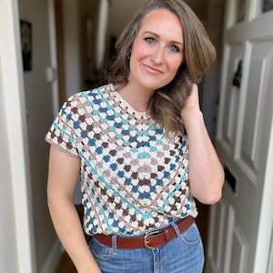 May include: A crocheted top with a geometric pattern in shades of brown, blue, and white. The top has short sleeves and a round neckline. The model is wearing a brown leather belt with a gold buckle.