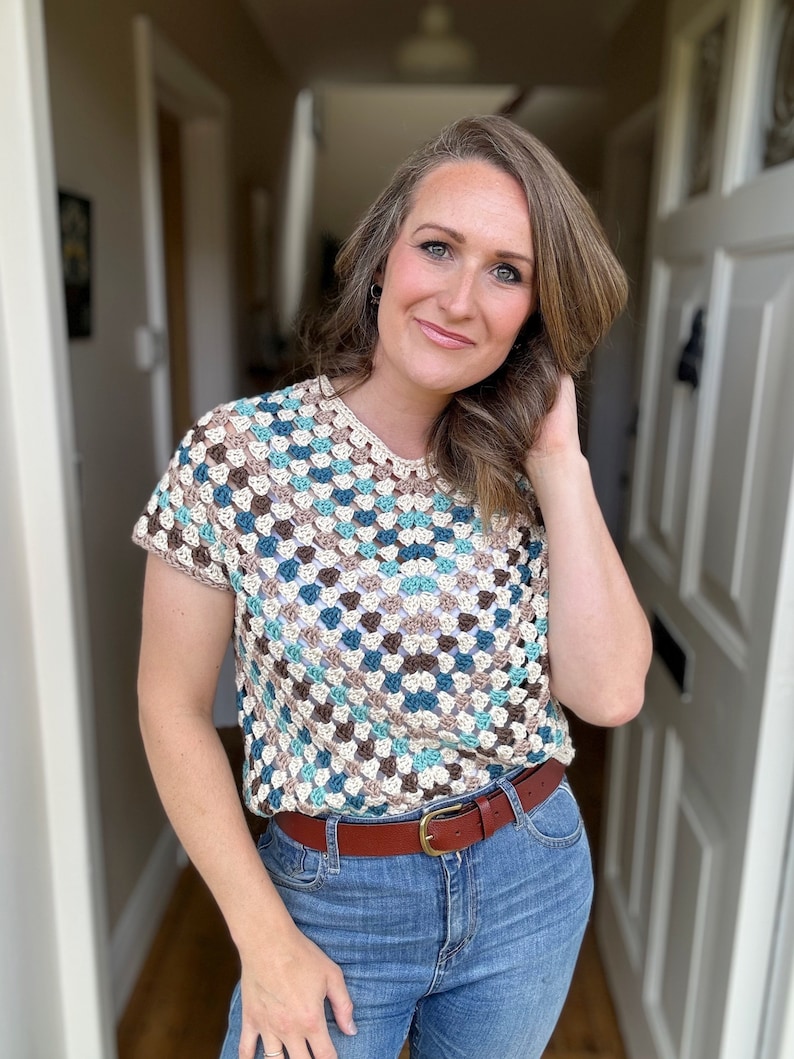 May include: A crocheted top with a geometric pattern in shades of brown, blue, and white. The top has short sleeves and a round neckline. The model is wearing a brown leather belt with a gold buckle.