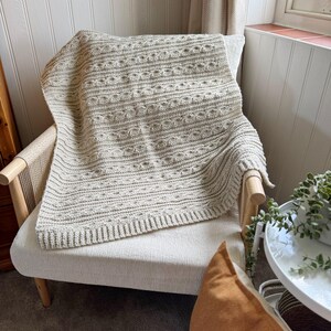 Peut inclure: Une couverture en tricot de couleur cr&egrave;me avec un motif textur&eacute; est drap&eacute;e sur un fauteuil de couleur claire. La couverture a une bordure c&ocirc;tel&eacute;e et un motif r&eacute;p&eacute;titif de lignes horizontales et de formes circulaires. La chaise a un cadre en bois et un dossier tiss&eacute;.