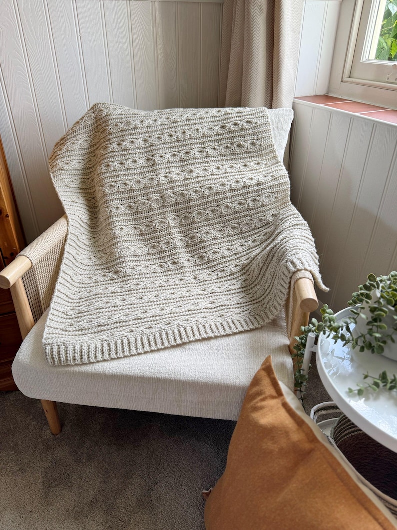 Peut inclure: Une couverture en tricot de couleur cr&egrave;me avec un motif textur&eacute; est drap&eacute;e sur un fauteuil de couleur claire. La couverture a une bordure c&ocirc;tel&eacute;e et un motif r&eacute;p&eacute;titif de lignes horizontales et de formes circulaires. La chaise a un cadre en bois et un dossier tiss&eacute;.