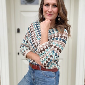 May include: A woman wearing a crocheted sweater with a brown, white, and blue geometric pattern. She is wearing blue jeans and a brown belt.