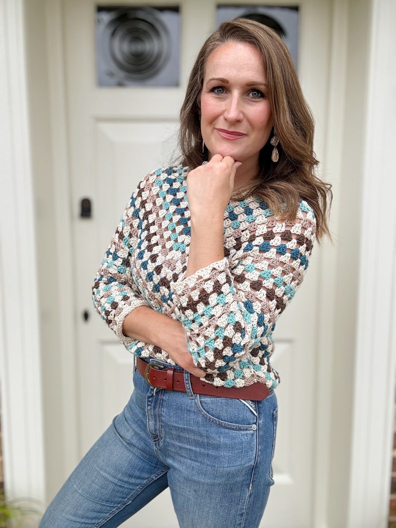 May include: A woman wearing a crocheted sweater with a brown, white, and blue geometric pattern. She is wearing blue jeans and a brown belt.