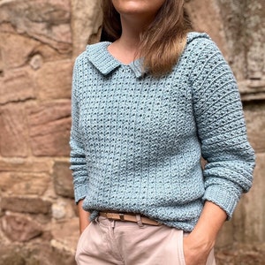 May include: A light blue crocheted sweater with a wide collar and long sleeves. The sweater has a textured stitch pattern and is worn with a brown belt.