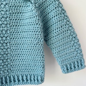 May include: A close-up of a light blue crocheted sweater with long sleeves. The sweater is made with a simple stitch pattern and has a ribbed hem and cuffs.