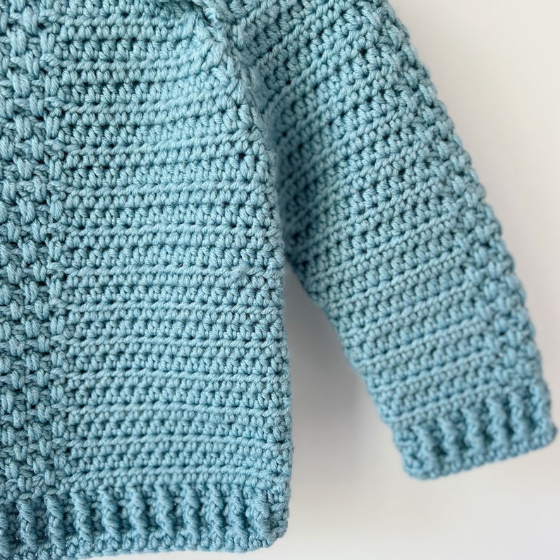 May include: A close-up of a light blue crocheted sweater with long sleeves. The sweater is made with a simple stitch pattern and has a ribbed hem and cuffs.