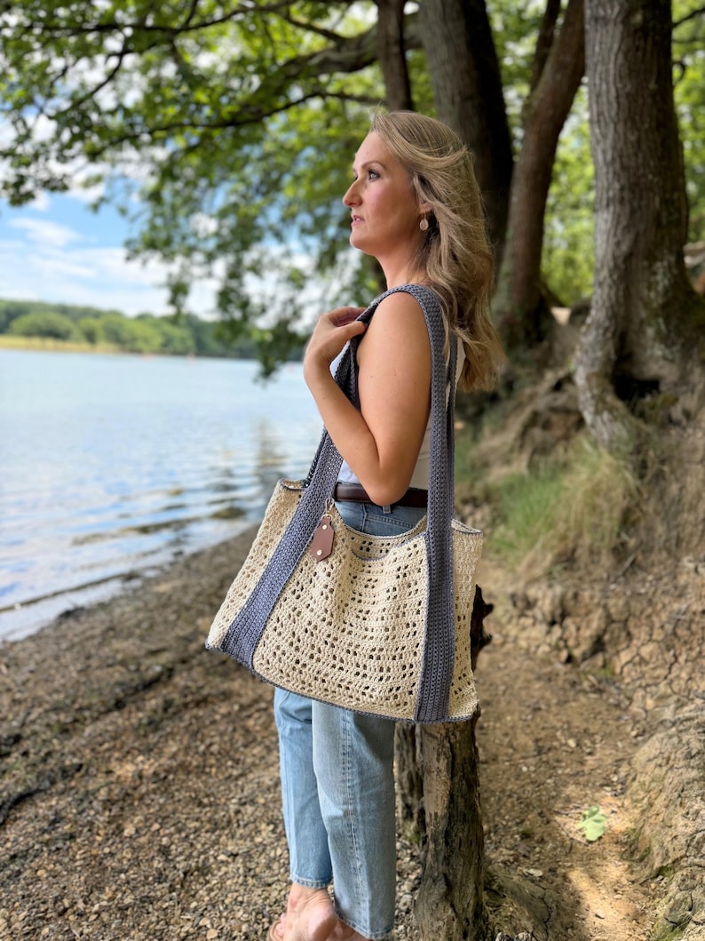 Puede incluir: Un bolso tote rectangular de ganchillo con un cuerpo calado de color crema y patr&oacute;n texturizado. El bolso incluye dos correas anchas de hombro de color gris oscuro y un borde gris oscuro, acentuado por una peque&ntilde;a etiqueta de cuero marr&oacute;n.