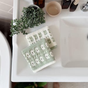 May include: Two handmade crochet washcloths with a floral pattern in a bathroom. The washcloths are white and light green. They are placed on a white sink, with a small plant, a bowl, and bottles.