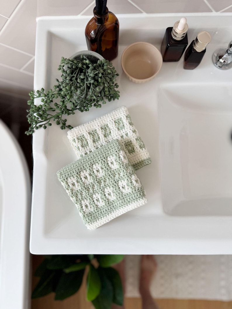 May include: Two handmade crochet washcloths with a floral pattern in a bathroom. The washcloths are white and light green. They are placed on a white sink, with a small plant, a bowl, and bottles.