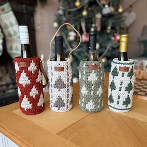 May include: Four wine bottles in decorative crochet bottle covers. The covers are in red, white, gray, and green, each with a Christmas tree pattern. The bottles sit on a wooden table, with a Christmas tree in the background.