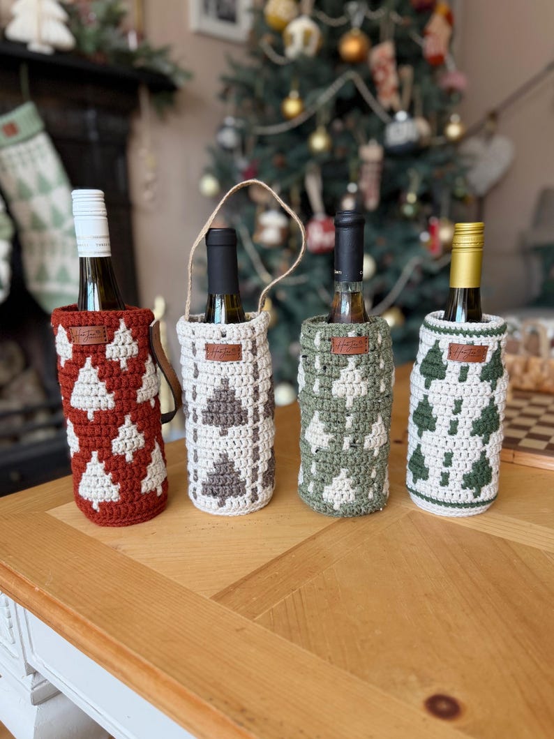 May include: Four wine bottles in decorative crochet bottle covers. The covers are in red, white, gray, and green, each with a Christmas tree pattern. The bottles sit on a wooden table, with a Christmas tree in the background.