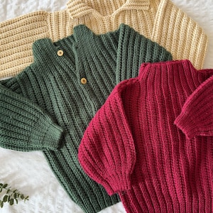 K&ouml;nnte beinhalten: Drei Strickpullover in verschiedenen Farben: hellbeige, dunkelgr&uuml;n und burgunderrot. Die Pullover liegen auf einer wei&szlig;en Oberfl&auml;che.