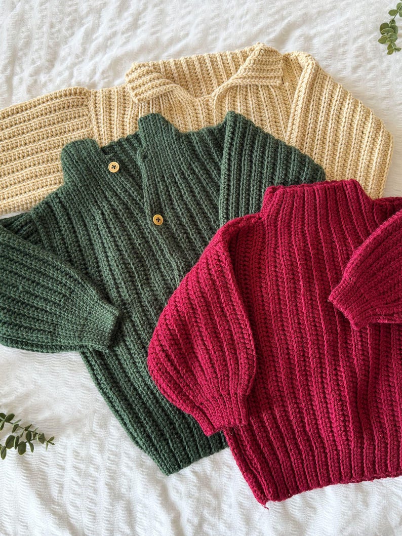 K&ouml;nnte beinhalten: Drei Strickpullover in verschiedenen Farben: hellbeige, dunkelgr&uuml;n und burgunderrot. Die Pullover liegen auf einer wei&szlig;en Oberfl&auml;che.