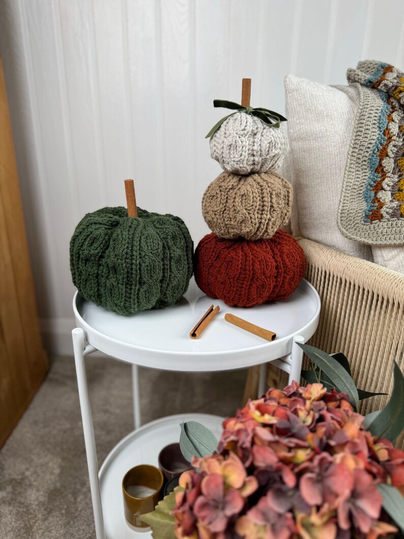May include: Four knitted pumpkins in autumnal colours: green, beige, and red, with cinnamon stick stems. They are stacked on a white, two-tiered side table. The top pumpkin has a green ribbon. A cozy, fall-themed decorative item for the home.