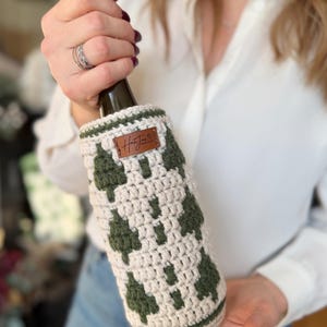 May include: Crocheted wine bottle cozy in cream and olive green. The cozy features a geometric pattern and a small brown leather tag with the text "HanJen". The cozy is designed to fit a standard wine bottle.
