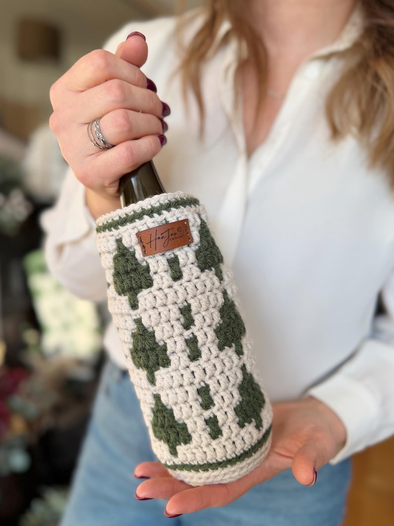 May include: Crocheted wine bottle cozy in cream and olive green. The cozy features a geometric pattern and a small brown leather tag with the text "HanJen". The cozy is designed to fit a standard wine bottle.