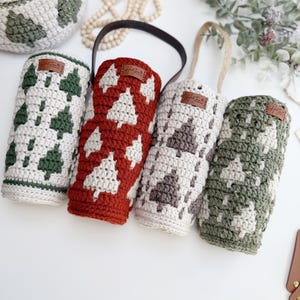 May include: Crocheted wine bottle holders in cream, red, gray, and green, each featuring a white Christmas tree design. The holders have leather tags and handles. A matching basket with a tree pattern is also visible.