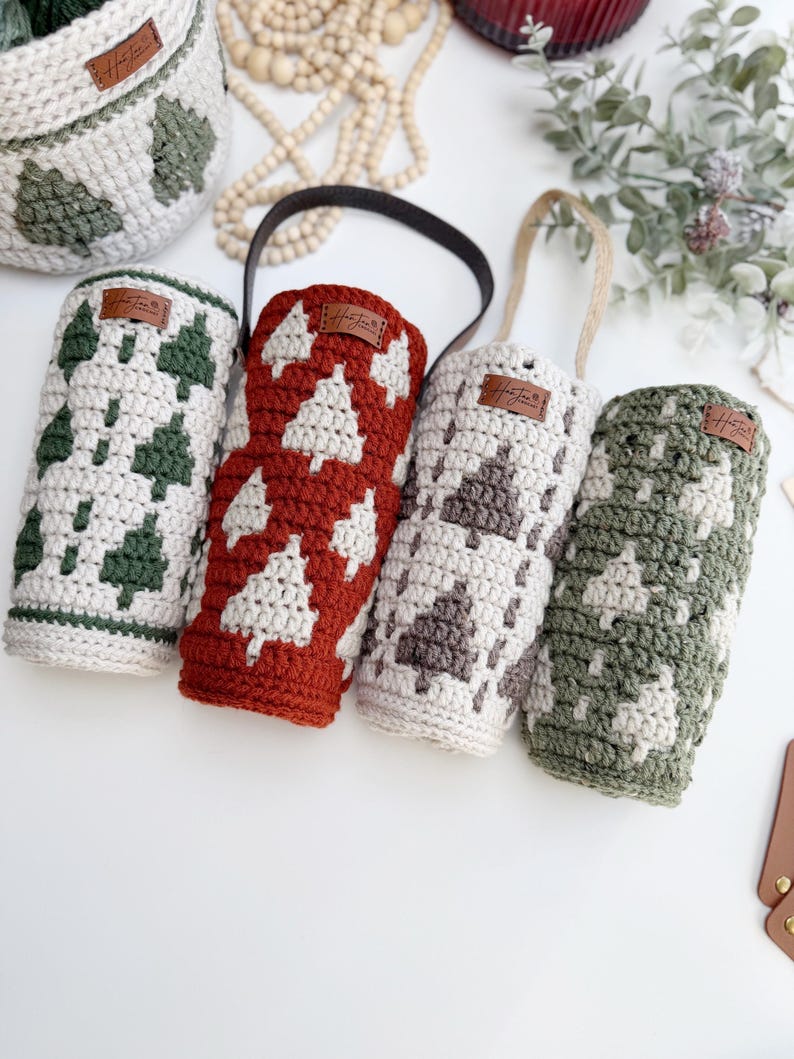 May include: Crocheted wine bottle holders in cream, red, gray, and green, each featuring a white Christmas tree design. The holders have leather tags and handles. A matching basket with a tree pattern is also visible.