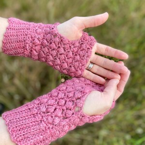 May include: A pair of pink crocheted fingerless gloves with a textured design. The gloves have a hole for the thumb.