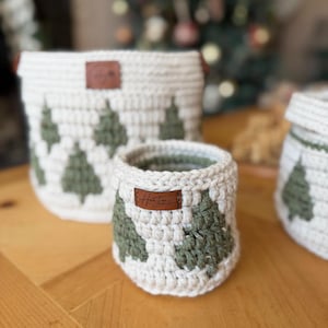 May include: Three handmade crochet baskets in varying sizes. The baskets are white with green Christmas tree designs. Each basket has a brown leather tag. The smallest basket has the text "HanJee Crochet" on the tag.
