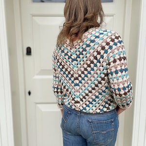 May include: A crocheted top with a geometric pattern in shades of brown, blue, and white. The top has a loose fit and three-quarter sleeves. The model is wearing blue jeans.