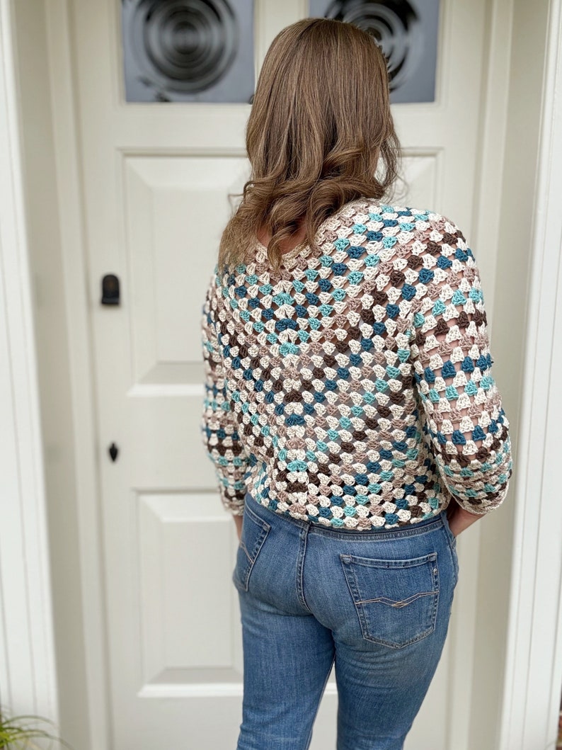 May include: A crocheted top with a geometric pattern in shades of brown, blue, and white. The top has a loose fit and three-quarter sleeves. The model is wearing blue jeans.