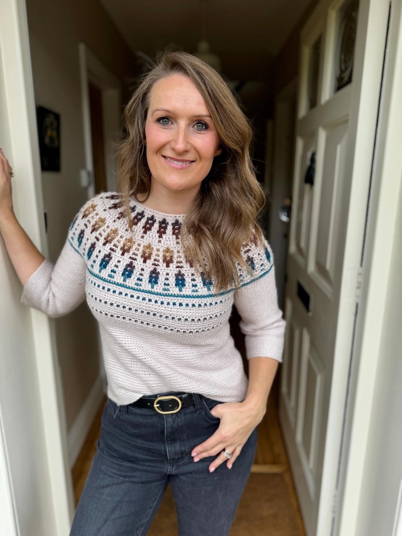 Pu&ograve; includere: Un maglione beige con uno sprone dettagliato con motivi geometrici marroni, verde acqua e bianchi. Il maglione ha maniche a tre quarti ed &egrave; abbinato a jeans scuri e una cintura nera con fibbia dorata. L'outfit &egrave; casual e confortevole.