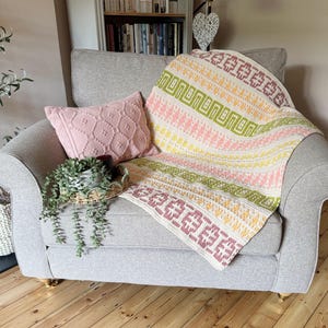 May include: A grey armchair with a colourful knitted blanket. The blanket has geometric patterns in pink, green, yellow, and purple. A pink cushion and a small plant in a basket are also visible.