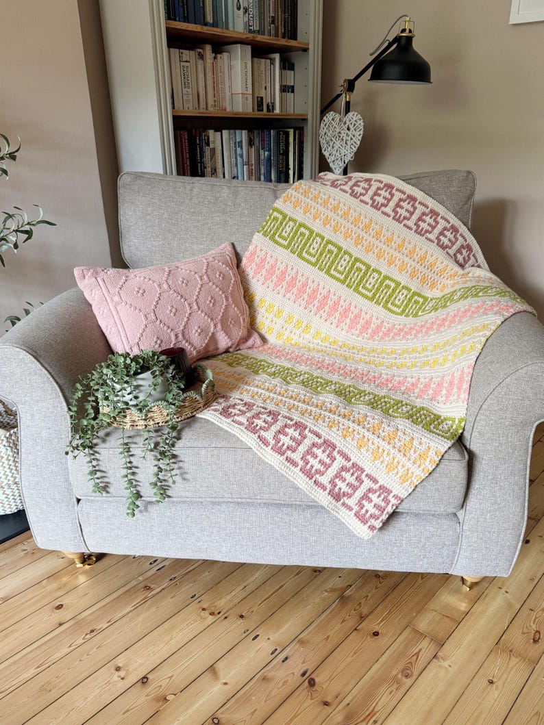 May include: A grey armchair with a colourful knitted blanket. The blanket has geometric patterns in pink, green, yellow, and purple. A pink cushion and a small plant in a basket are also visible.