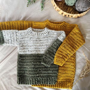 Puede incluir: Dos su&eacute;teres de crochet, uno de color amarillo mostaza y el otro con un patr&oacute;n de rayas multicolores de blanco, gris y verde.
