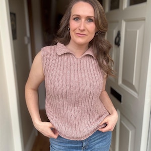 May include: A sleeveless, collared, dusty rose-coloured knit top. The top has a ribbed texture and a V-neck collar. The person in the image is wearing blue jeans. The background is a doorway.