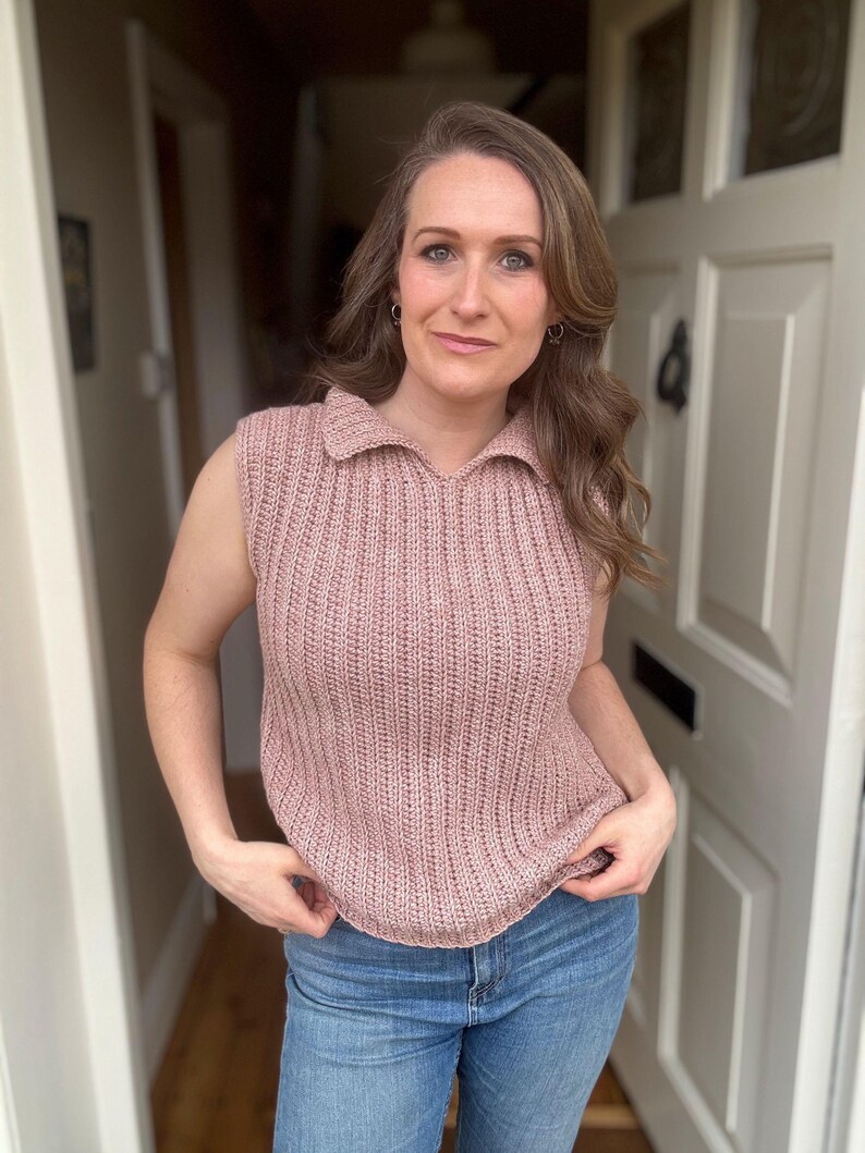May include: A sleeveless, collared, dusty rose-coloured knit top. The top has a ribbed texture and a V-neck collar. The person in the image is wearing blue jeans. The background is a doorway.