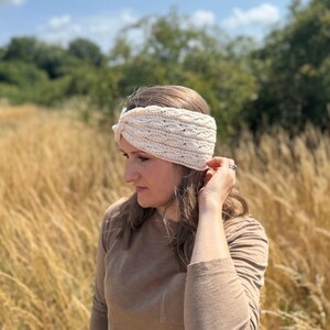 May include: Cream-colored knitted headband with a textured design. The headband is worn on the head and has a twisted knot detail. The image is taken outdoors with a background of tall grass and trees.