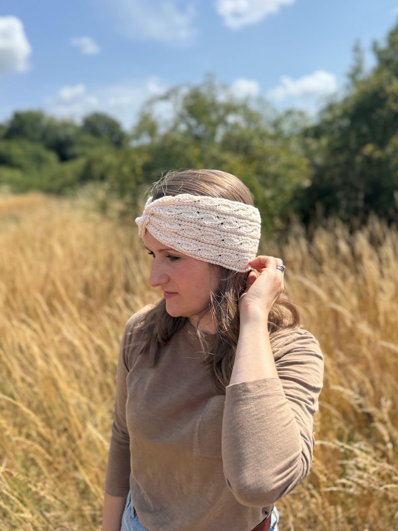 May include: Cream-colored knitted headband with a textured design. The headband is worn on the head and has a twisted knot detail. The image is taken outdoors with a background of tall grass and trees.
