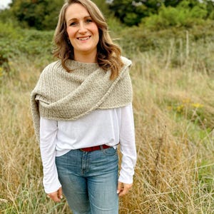 May include: A woman wearing a white long-sleeved shirt, a brown knitted shawl, and blue jeans. She is standing in a field of tall grass.