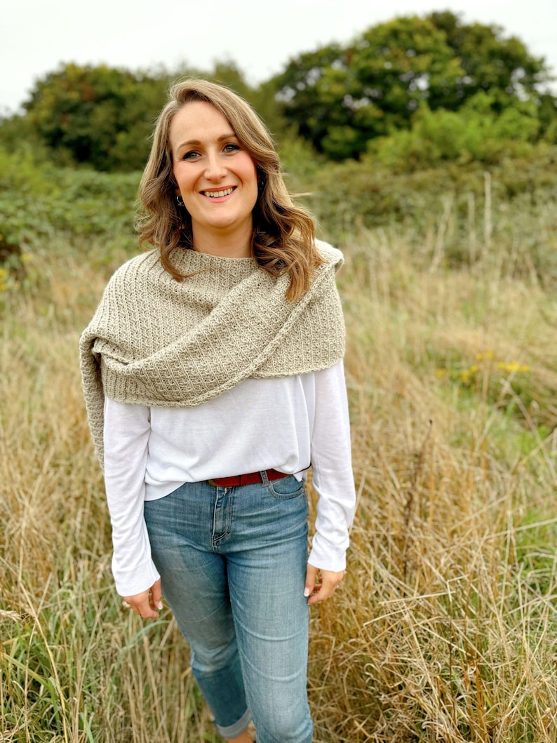 May include: A woman wearing a white long-sleeved shirt, a brown knitted shawl, and blue jeans. She is standing in a field of tall grass.
